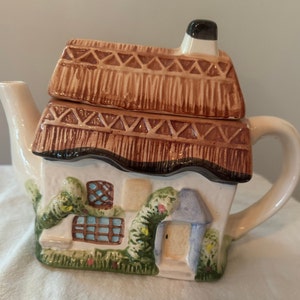 Peut inclure: Une théière en céramique blanche en forme de cottage avec un toit de chaume brun, du lierre vert et des fenêtres bleues et blanches. La théière a un bec verseur et une poignée.