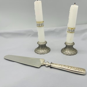 May include: Two white candles with decorative rhinestone bands in ornate silver candle holders. A silver cake server with a detailed handle lies in front. The items are on a light grey surface.
