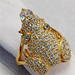 May include: A gold-colored ring in the shape of a frog, encrusted with numerous clear, sparkling gemstones. The frog's body is covered in the gems, with larger stones for the eyes and a prominent stone on its head. The ring has a detailed, textured design.