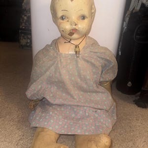 May include: An antique doll with a painted face, wearing a pink floral dress and a necklace. The doll shows signs of wear and tear. The doll is sitting on a carpeted surface.