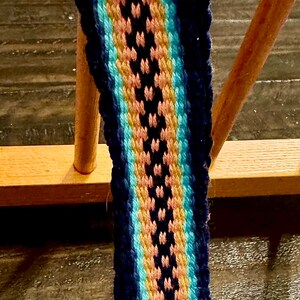 May include: A woven strap with a repeating pattern of pink, yellow, and blue stripes against a navy blue background. The strap has a silver-colored metal clasp with a D-ring.