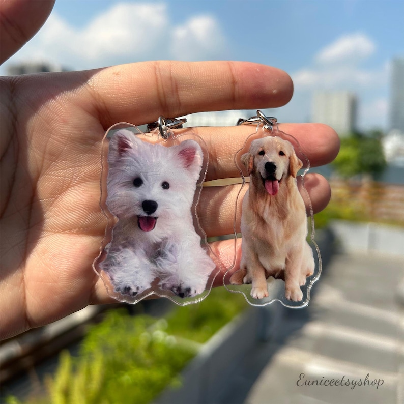 May include: Two clear acrylic keychains with images of dogs. One keychain features a white West Highland White Terrier, and the other keychain features a golden retriever.