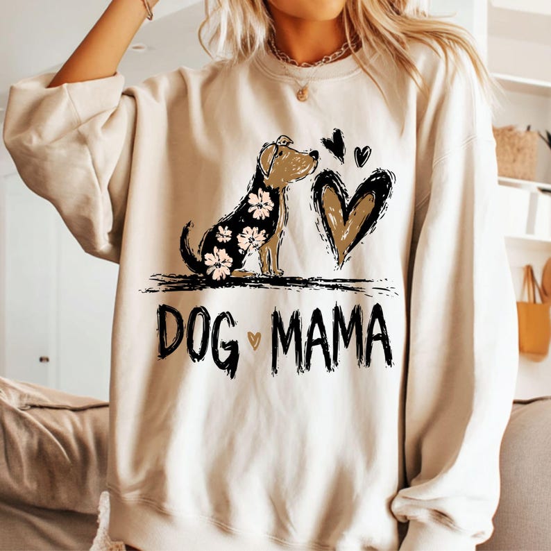 May include: A cream-coloured sweatshirt with a graphic of a dog with floral accents, a heart, and the text "DOG MAMA" in black. The dog has a black body with white flowers. The heart is brown with a black outline.