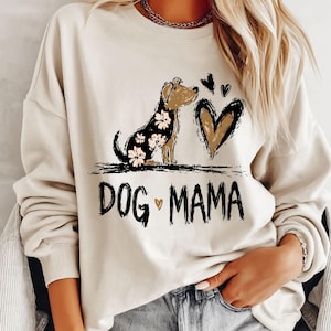 May include: A cream-coloured sweatshirt with a dog illustration, floral accents, and a heart design. The text "DOG MAMA" is printed in black. A casual, comfortable top for dog lovers.