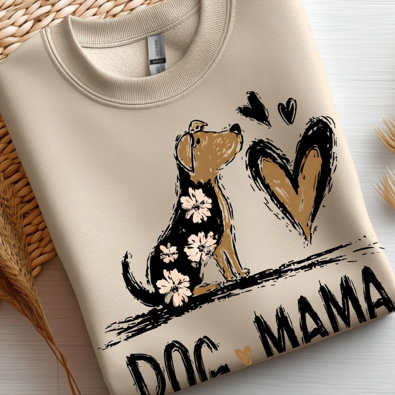 May include: Beige sweatshirt featuring a brown and black dog illustration with floral details, a heart, and the text "DOG MAMA". The dog is looking up towards the hearts. The sweatshirt is made of a soft material.