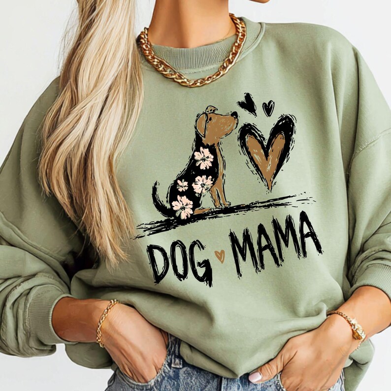 May include: Sage green sweatshirt featuring a dog graphic with floral details and a heart. The words "DOG MAMA" are printed in black. A gold chain necklace and bracelet are visible.