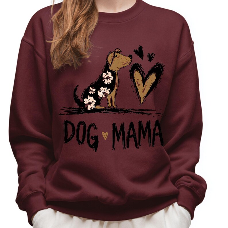 May include: Burgundy sweatshirt featuring a dog graphic with white floral accents, a heart, and the text "DOG MAMA". The dog is brown and black. The heart is brown with a black outline. The text is black.