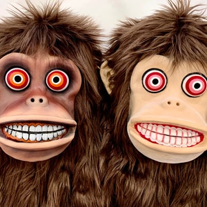 May include: Two brown furry monkey masks with red eyes and white teeth. The eyes have concentric circles of orange and white.