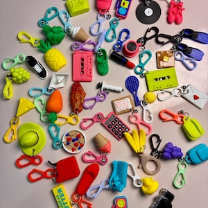 May include: A collection of colorful miniature plastic keychains featuring various shapes including food items like a pineapple, carrot, and butter stick, along with objects such as a cassette tape, boombox, calculator, sneakers, and sports equipment.