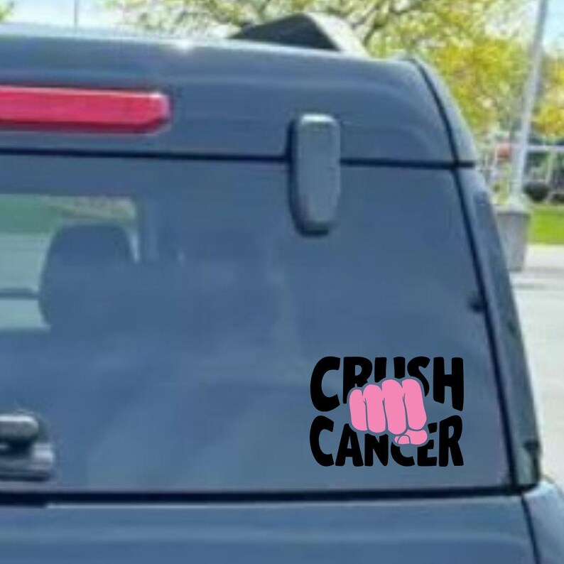 May include: Black decal with the words "CRUSH CANCER" and a pink fist graphic. The decal is affixed to the rear window of a dark blue vehicle.