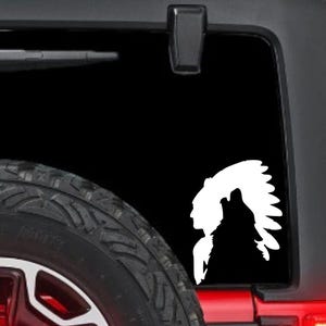 May include: White vinyl decal featuring a Native American profile and a howling wolf silhouette. The decal is applied to a black vehicle window, with a portion of the tire visible in the foreground.