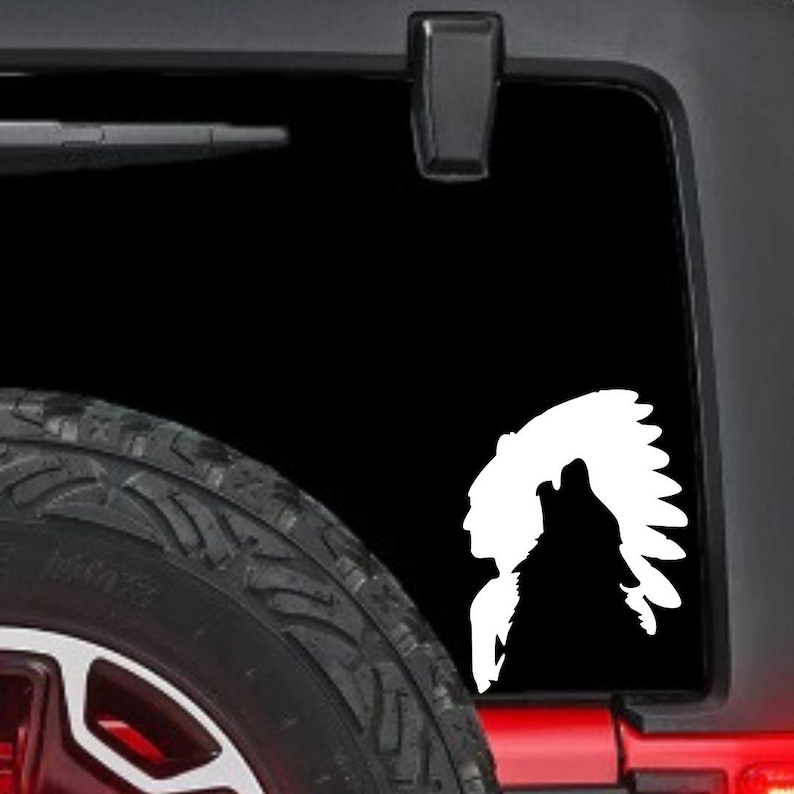 May include: White vinyl decal featuring a Native American profile and a howling wolf silhouette. The decal is applied to a black vehicle window, with a portion of the tire visible in the foreground.