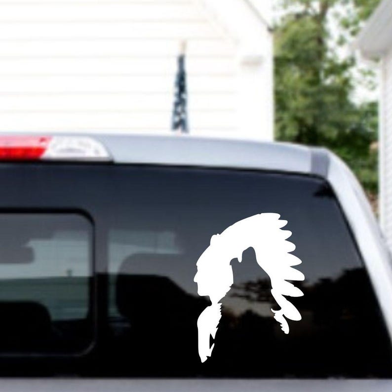 May include: White vinyl decal of a Native American chief in profile, wearing a feathered headdress. The decal is applied to the rear window of a vehicle. The design is a silhouette, with the details of the face and headdress clearly visible.