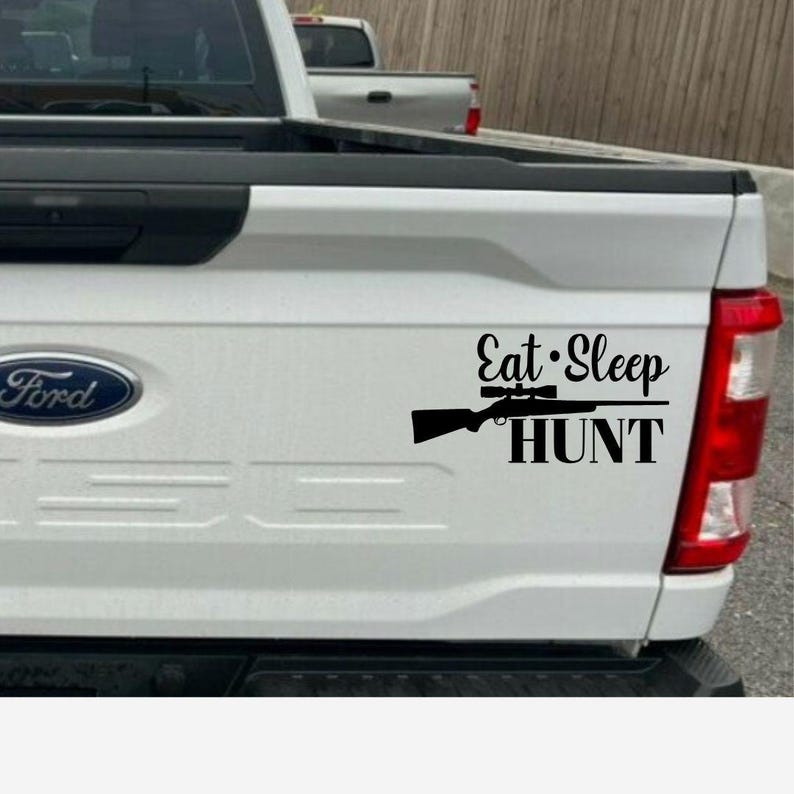 May include: Black vinyl decal on a white truck tailgate. The decal features the words "Eat Sleep HUNT" with a rifle graphic. The Ford logo is visible on the left side of the tailgate.