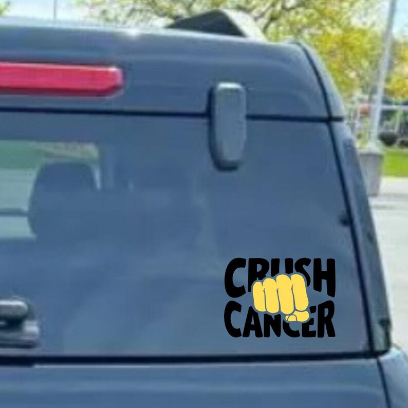 May include: Black and yellow "Crush Cancer" decal on a car window. The text is in a bold, sans-serif font, with the word "Crush" above "Cancer". A yellow fist graphic is between the words.