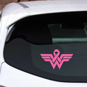 May include: Pink Wonder Woman logo with a pink ribbon in the center. The logo is on a black car window.