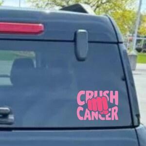 May include: Pink decal with the words "CRUSH CANCER" and a fist graphic. The decal is displayed on the rear window of a dark-colored vehicle. The text and fist are in a bold, sans-serif font.