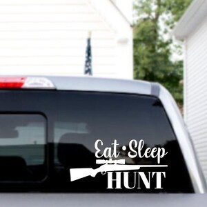 May include: White vinyl decal with the words "Eat Sleep HUNT" and a rifle graphic. The decal is applied to the rear window of a vehicle, with a red brake light visible above.