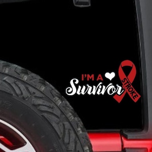 May include: A black decal with the words "I'M A Survivor" in white script, a white heart, and a red ribbon with the word "STROKE". The decal is on a black surface, with a tire visible in the foreground.