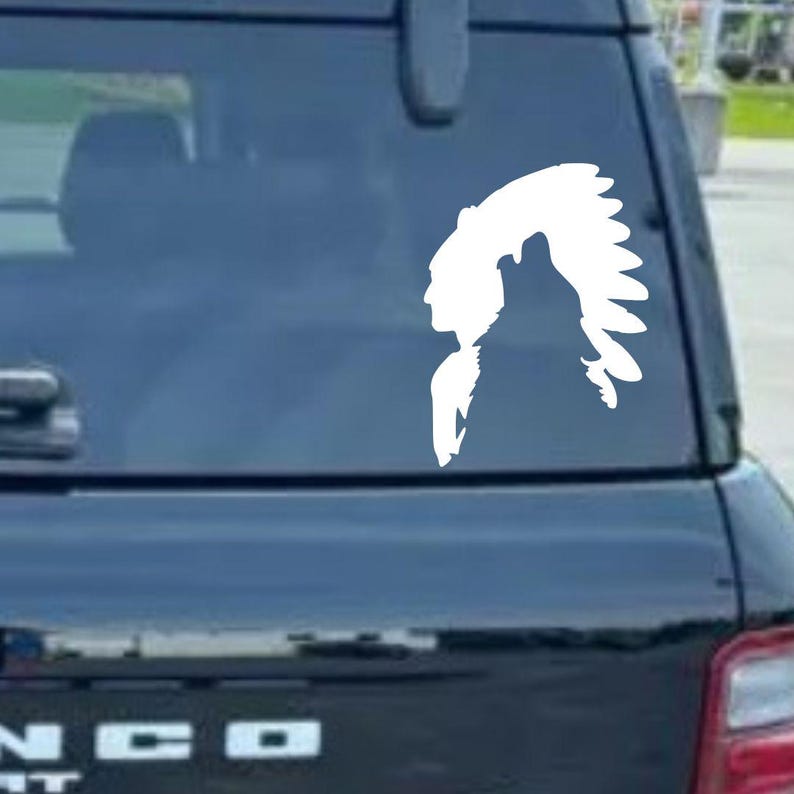 May include: White vinyl decal featuring a Native American profile with a wolf howling. The design is a silhouette, suitable for application on a car window or other smooth surfaces. The decal is a single color and appears to be die-cut.