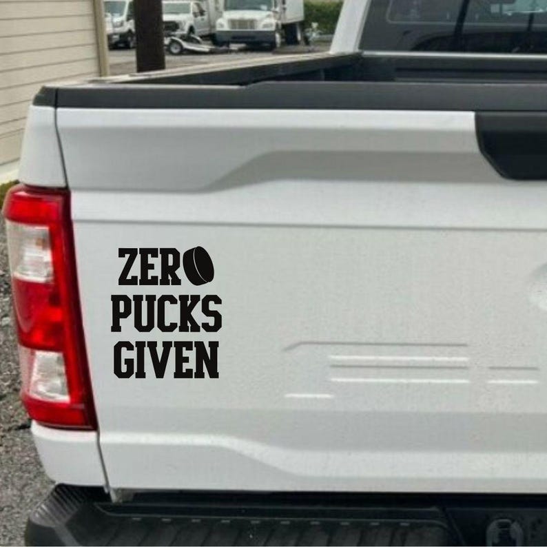 May include: A black decal with the words "ZERO PUCKS GIVEN" and a hockey puck graphic is affixed to the white tailgate of a truck. The text is in a bold, sans-serif font.