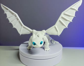 May include: White 3D printed figurine of a dragon with blue eyes. The dragon is posed with its wings spread out and its head tilted down. The figurine is sitting on a white circular base.