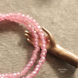 May include: A pink beaded necklace with a gold hand charm. The charm has the text "... arte bella ... 10727" on it.