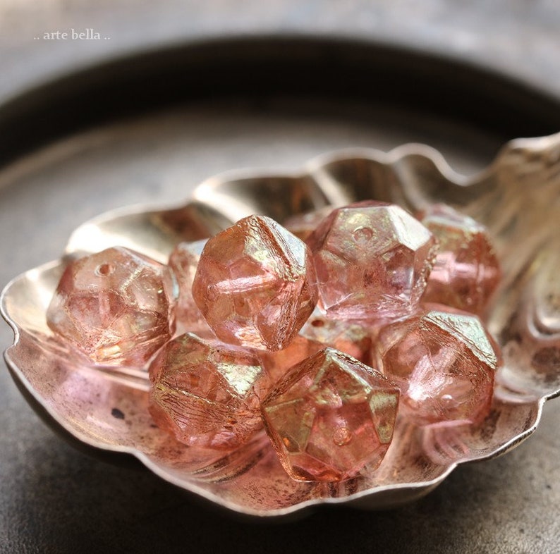 Op de afbeelding: Close-up van gefacetteerde, roze kralen met iriserende highlights, rustend in een zilveren, bladvormige schaal. De kralen hebben een geometrische, kristalachtige uitstraling. De afbeelding is zacht belicht, waardoor de texturen en kleuren van de kralen worden benadrukt.