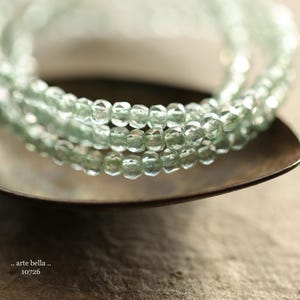 May include: A close-up of a multi-strand necklace made of light green faceted beads. The necklace is lying on a brown surface.