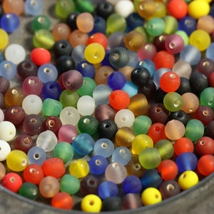 May include: A bowl filled with a variety of colorful glass beads. The beads are opaque and have a frosted finish. They are in a range of colors, including red, blue, green, yellow, and purple.