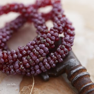 May include: Close-up of a deep red beaded necklace, intricately woven. The beads have a textured, clustered appearance. The necklace is draped over a vintage, rusty key, with the text ".. arte bella .." in the lower left corner.