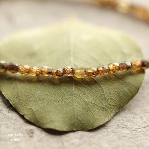May include: A delicate necklace featuring small, faceted amber-colored beads. The beads are strung together, resting on a green leaf. The background is a textured, neutral surface. The text "..arte bella.. 11068" is visible in the lower left corner.