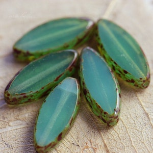 May include: Close-up of six oval-shaped glass beads. The beads are a translucent teal color with a green stripe down the center. The edges are a speckled brown and green. The beads are arranged on a light brown surface.