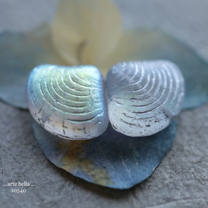 May include: Two iridescent blue and green glass shell-shaped beads with a textured surface. The beads are on a blue and white background.