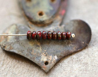 May include: A strand of eight dark red and black oval beads on a silver pin. The beads are arranged on a rustic, heart-shaped metal object. The background features a blurred, textured surface.
