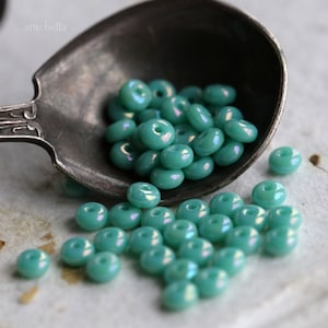 May include: A collection of turquoise iridescent glass beads in a silver spoon. The beads are round with a hole in the center.