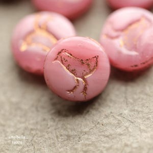 May include: Close-up of several pink, round beads with a gold-colored bird design. The beads have a glossy finish and are arranged on a textured, gray surface. The text "arte bella" and "11074" is visible.