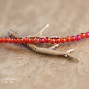 May include: A strand of small, round, iridescent red beads arranged in a horizontal line, resting across a silver-toned, textured, branch-shaped metal object. The text ".. arte bella .. 11613" is printed in the bottom left corner of the image.