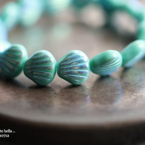 May include: A strand of iridescent teal blue glass beads in the shape of seashells. The beads are strung together to form a bracelet.