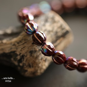 May include: A close-up of a dark red iridescent beaded bracelet. The beads are faceted and have a subtle shimmer. The bracelet is lying on a piece of weathered wood.