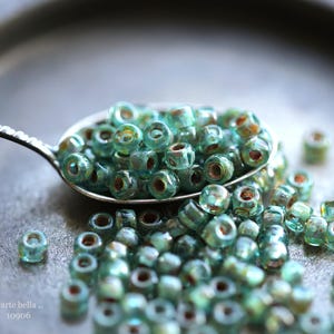 May include: A close-up of a spoon filled with teal and green glass beads. The beads are scattered on a gray surface.  ...arte bella... 10906