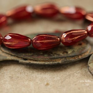 May include: A strand of faceted, teardrop-shaped beads in a deep, translucent red hue with metallic copper-colored accents. The beads are arranged in a circular formation. The text 'arte bella .. 11549' is visible in the lower right corner.