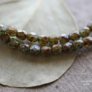 May include: A strand of green and brown glass beads with a mottled, textured surface. The beads are arranged in a circular pattern and are strung on a clear thread.