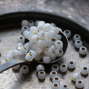May include: A close-up of a pile of white glass beads with gold accents. The beads are in a silver spoon on a gray metal surface.