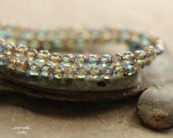 May include: Two strands of iridescent beaded bracelet. The beads are a mix of yellow, blue, and clear, strung together with a neutral-colored cord. The bracelet is resting on a textured, weathered surface. The text "arte bella" and "11085" is visible.