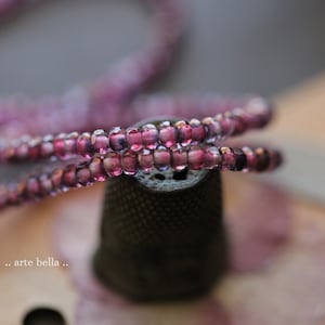 May include: A close-up of a strand of purple glass beads wrapped around a small, dark gray thimble. The beads are translucent and have a slight shimmer.