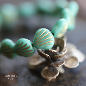 May include: A close-up of a teal blue and gold beaded necklace. The beads are shaped like seashells and have a sunburst design. The necklace is resting on a small, silver, flower-shaped stand.