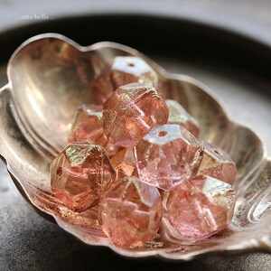 Op de afbeelding: Close-up van een verzameling gefacetteerde, roze kralen met een metallic glans. De kralen liggen in een decoratief, zilverkleurig schaaltje. De kralen hebben een geometrische vorm en zijn waarschijnlijk bedoeld voor het maken van sieraden of knutselen.