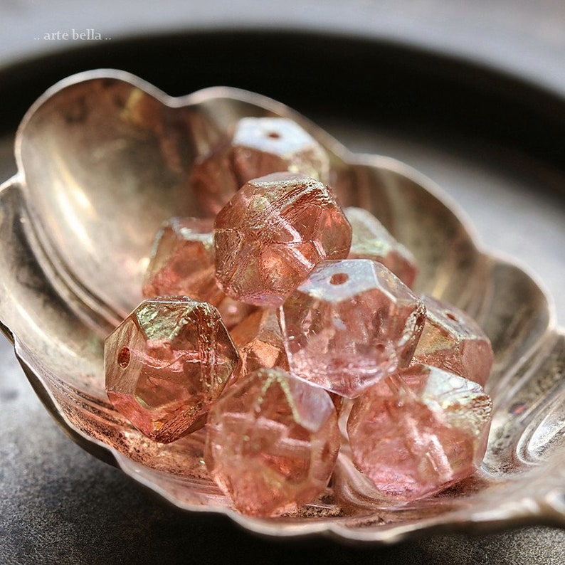 Op de afbeelding: Close-up van een verzameling gefacetteerde, roze kralen met een metallic glans. De kralen liggen in een decoratief, zilverkleurig schaaltje. De kralen hebben een geometrische vorm en zijn waarschijnlijk bedoeld voor het maken van sieraden of knutselen.