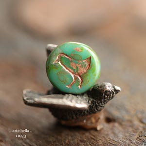 May include: A handcrafted bead with a teal green hue and a copper-colored bird design. The bead is mounted on a small, detailed silver-toned bird figurine. The image is a close-up, showcasing the bead's texture and the figurine's intricate details.